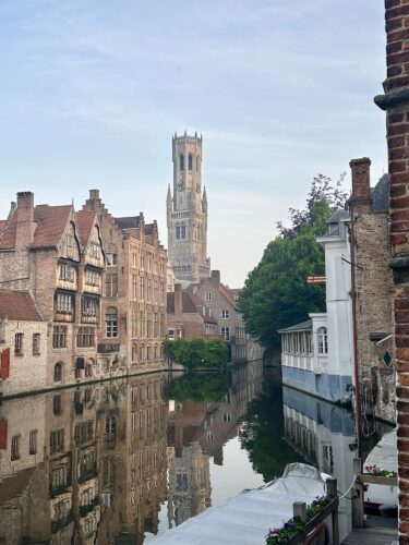 View of a historic canal in Bruges, Belgium, featuring traditional brick buildings and a tall church tower reflected in the water, under a clear sky.