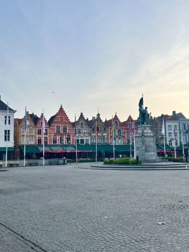 A peaceful square in Bruges, featuring historic buildings with colorful facades and a central statue, set against a clear sky.