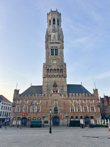 Historic belfry tower and municipal building in a town square, featuring gothic architecture and a clock.