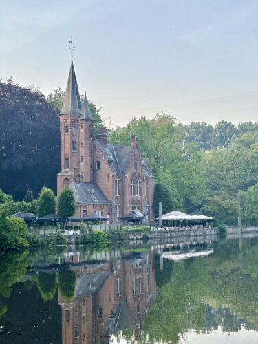 A picturesque brick castle with a tall spire, surrounded by green foliage, reflecting in a calm water body.