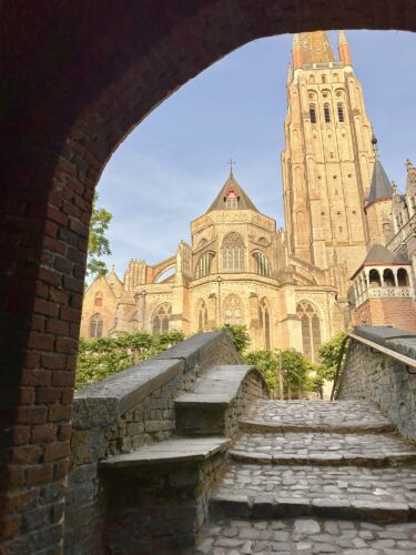 View of a historic church towering above a stone staircase, framed by an archway with greenery in the foreground.