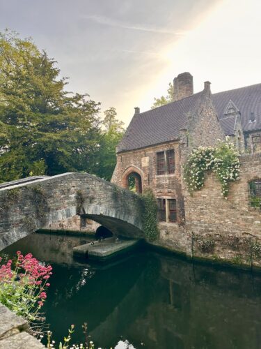 A picturesque stone bridge arching over a calm canal, with a historic building adorned with greenery and flowers in the background, set against a serene sky.