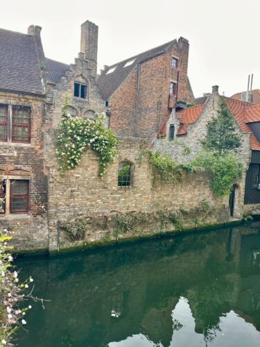 Historic brick buildings alongside a tranquil canal, with greenery and flowers growing on a stone wall.