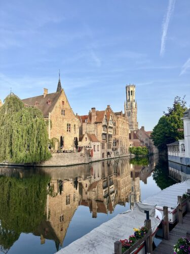 Scenic view of medieval buildings along a canal in Bruges, Belgium, with a clear blue sky and reflections in the water.
