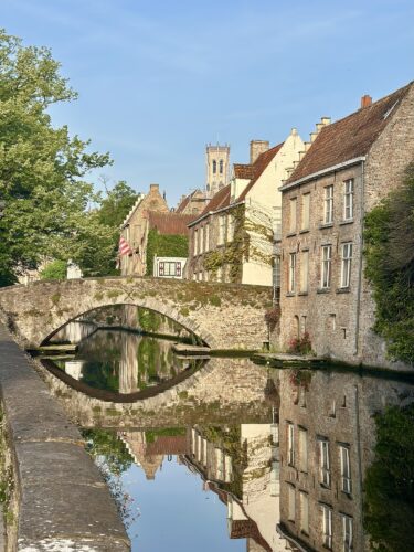 A picturesque canal scene featuring a stone bridge, surrounded by historic buildings and trees, reflecting in the calm water below.