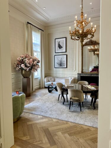An elegantly decorated room featuring a chandelier, large floral arrangement, framed artworks on the wall, and a dining table surrounded by stylish chairs.