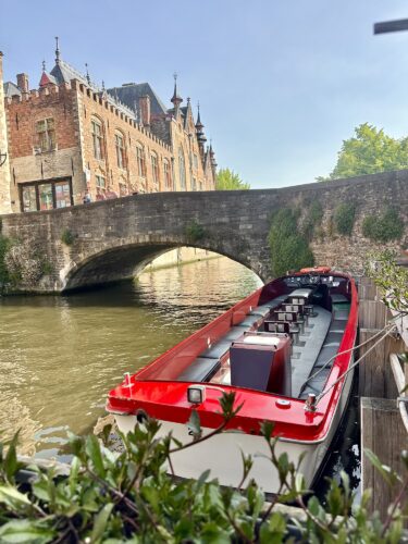 A red boat docked along a canal beneath a stone bridge, with historic buildings in the background under a clear sky.