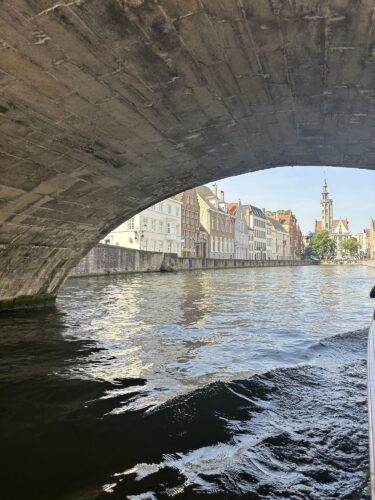 View from under a stone bridge overlooking a river with historic buildings on the banks and a church steeple in the distance.