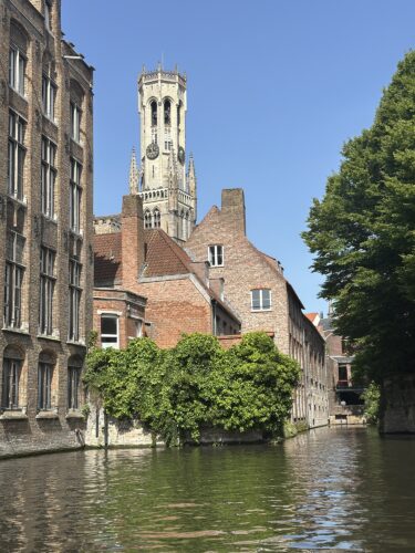 A scenic view of a canal in Bruges, Belgium, featuring historic brick buildings and a medieval bell tower under a clear blue sky.