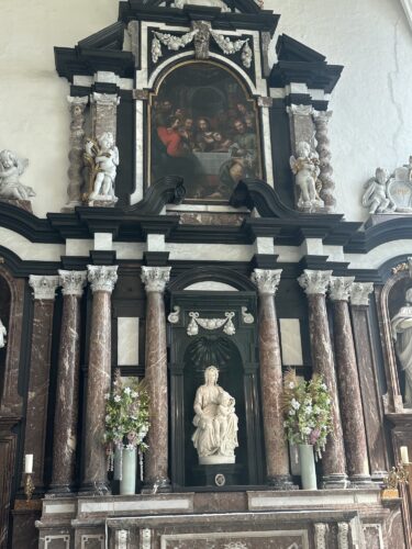 A richly decorated altar featuring marble columns, a statue of a figure at the center, and a painting depicting a group of people in the background. Floral arrangements complement the overall design.