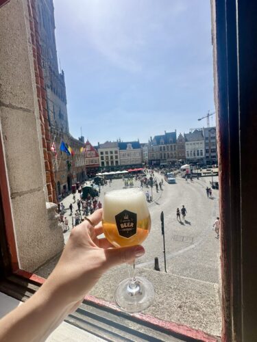 A hand holding a glass of beer in front of a window with a view of a busy square, featuring historic buildings and people walking around.