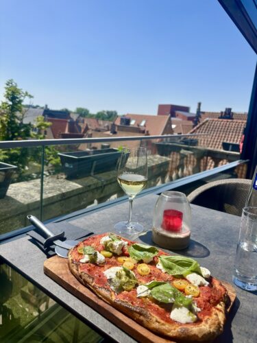 A freshly made pizza topped with tomatoes, mozzarella, basil, and pesto on a wooden board, accompanied by a glass of white wine, set on a balcony with a view of rooftops and a clear blue sky.