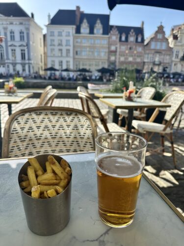 A glass of beer and a metal cup of fries on a table overlooking a canal with historic buildings in the background.