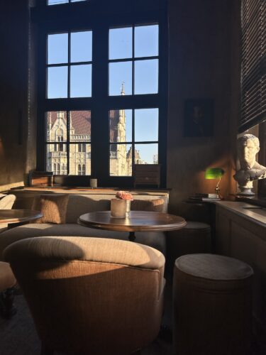 Cozy interior of a cafe with soft armchairs and round tables, featuring large windows that offer a view of an ornate building outside, illuminated by natural sunlight.
