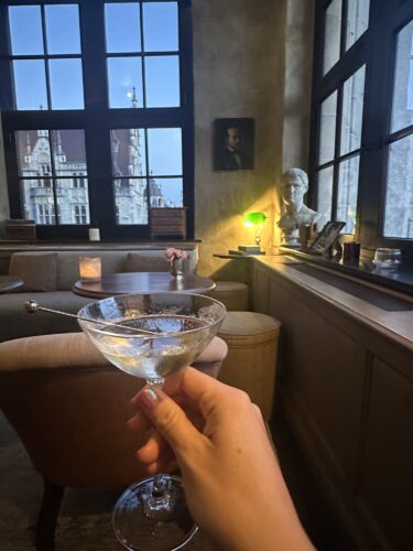 A hand holding a cocktail glass in a cozy, well-lit lounge with large windows showcasing an outdoor view, including a statue and a portrait on the wall.