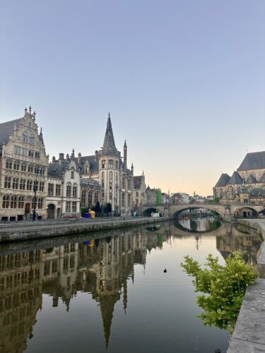 View of historic buildings along a river in Ghent, Belgium, with reflections in the water and a clear sky.