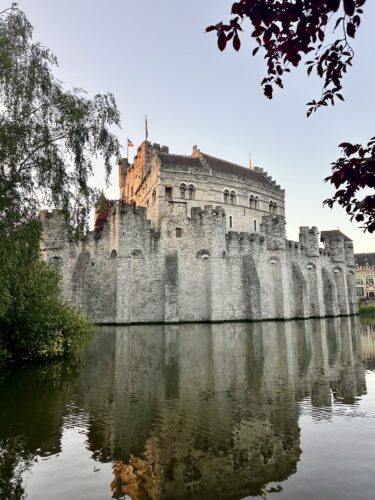 A medieval castle surrounded by water, featuring tall stone walls and turrets, reflecting in the calm surface of the lake during sunset.
