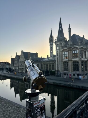 A telescope overlooking a canal in Ghent, Belgium, with historic buildings and a clear sky in the background.