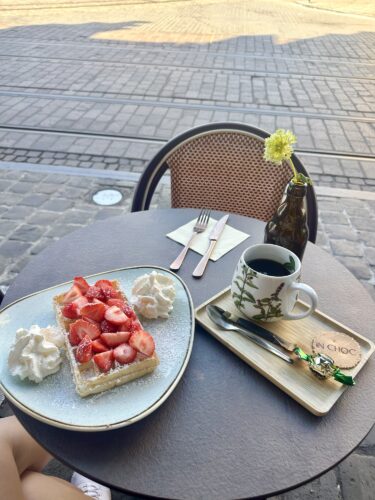 A table set for a meal outdoors featuring a slice of strawberry cake topped with whipped cream, a cup of coffee, and a small bottle with a flower, against the backdrop of a cobblestone street.