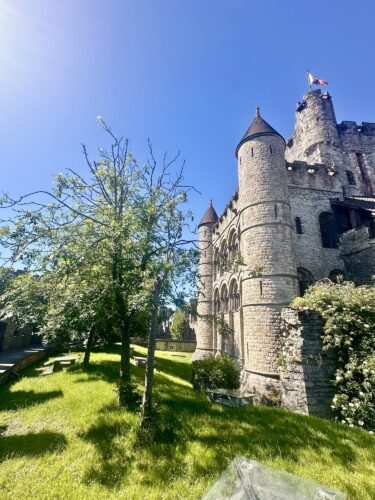 A stone castle with turrets, surrounded by green grass and trees under a clear blue sky.