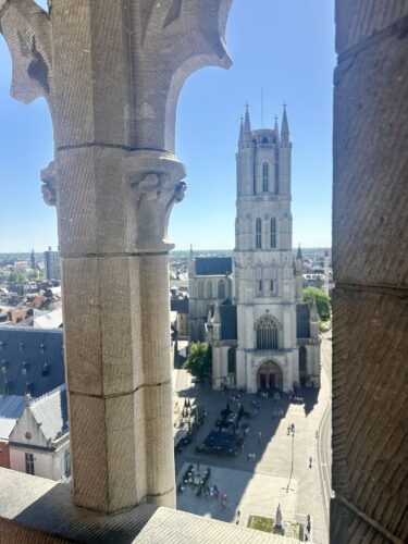 View of a tall bell tower and a church from a stone window, with a plaza and people below in a sunny setting.