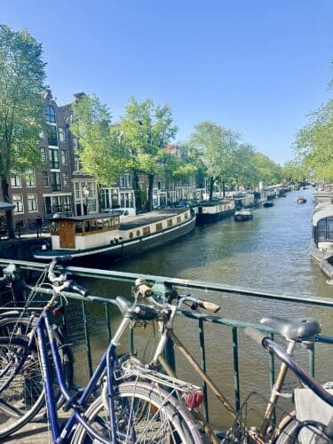Scenic view of a canal in Amsterdam with green trees lining the water, boats moored alongside the banks, and bicycles parked on a bridge in the foreground.
