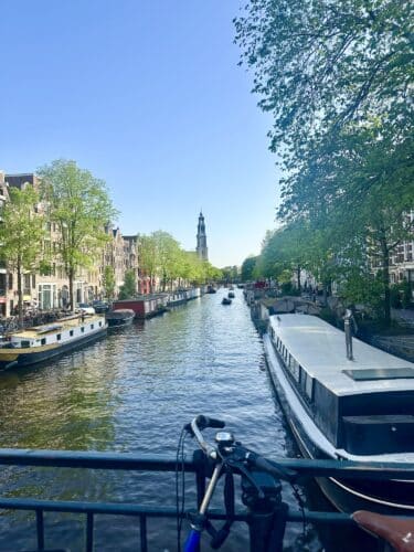 View of a canal in Amsterdam with boats and lush green trees lining the banks, featuring a tower in the background under a clear blue sky.