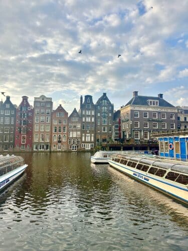 View of colorful historic buildings along a canal in Amsterdam, with boats moored in the foreground and a cloudy sky above.