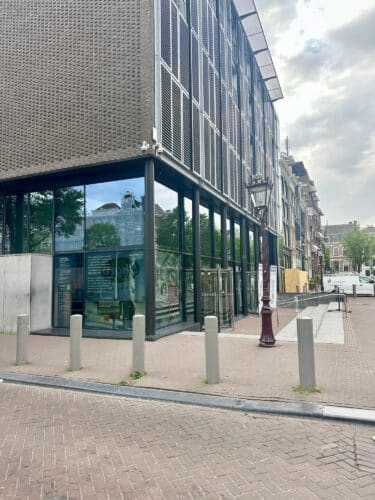 Modern building with glass windows and metal mesh facade, situated on a brick-paved street lined with bollards and greenery.