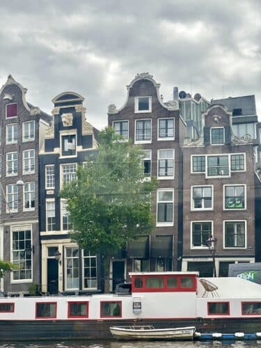 Row of traditional Dutch canal houses with gabled roofs in Amsterdam, alongside a boat moored on the water, under a cloudy sky.
