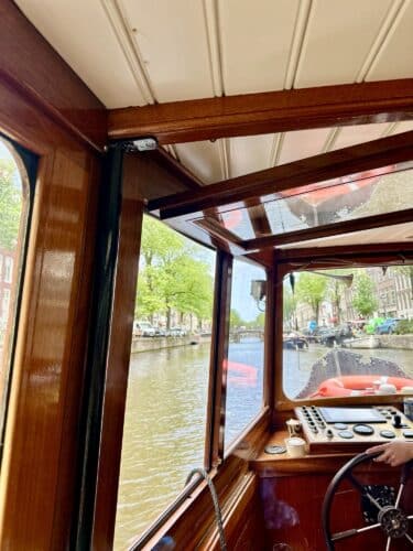Interior view of a boat navigating a canal, featuring wooden panels and a steering wheel, with greenery and buildings visible through the windows.