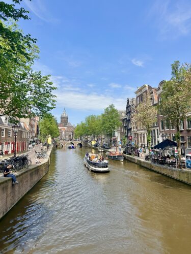 A picturesque view of a canal in Amsterdam with boats, surrounded by vibrant trees and historic buildings under a clear blue sky.