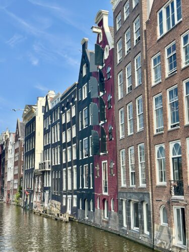 Row of traditional Dutch canal houses in Amsterdam, featuring distinct architectural styles and colors, with a clear blue sky reflected in the water of the canal.