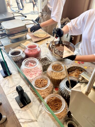 A dessert shop counter featuring jars of colorful toppings such as sprinkles, nuts, and candies, with an employee preparing a treat in the foreground.