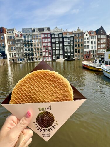 A person holding a Stroopwafel from the 'Strooperie' in front of a picturesque canal lined with colorful buildings in Amsterdam.