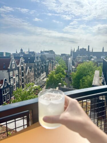 A hand holding a glass of beverage in front of a scenic view of Amsterdam's architecture, featuring tall buildings and green trees under a bright sky.