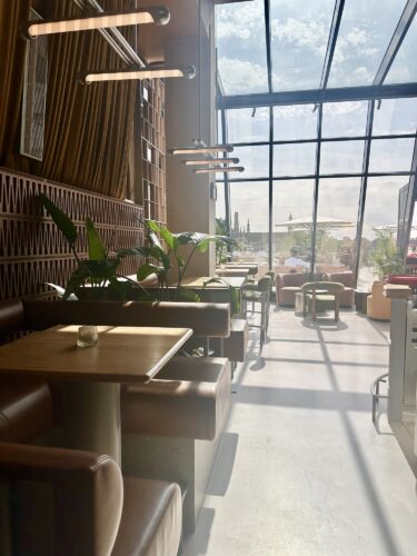 Interior of a modern café with large glass windows, featuring tables and chairs, plants, and pendant lights casting shadows on the floor.
