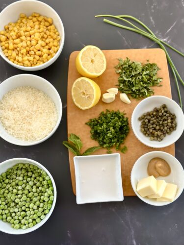 Various ingredients arranged on a wooden cutting board, including yellow corn, lemon halves, garlic, fresh herbs, green peas, rice, capers, a bowl of yogurt, mustard, and butter.