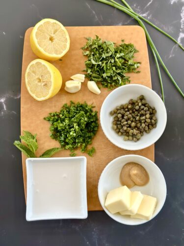 A wooden cutting board displaying various ingredients for cooking: halved lemons, chopped herbs (mint and cilantro), garlic cloves, capers in a bowl, a bowl of tahini, a small bowl of mustard, and cubes of butter. Green chives are visible in the background.