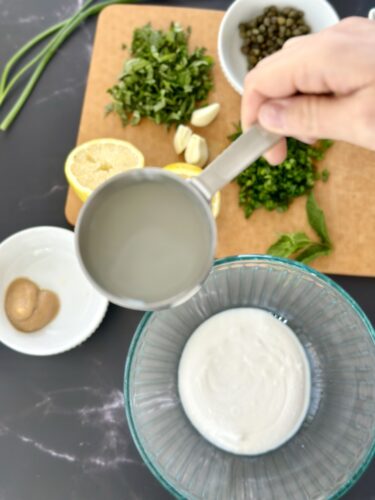 A person pouring liquid from a measuring cup into a bowl containing a creamy substance, surrounded by fresh herbs, garlic, lemon, capers, and a small dish with a brown condiment on a wooden cutting board.