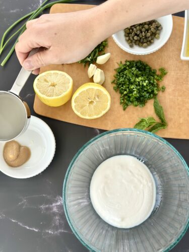 A hand pouring liquid into a bowl, surrounded by fresh ingredients including lemon halves, garlic cloves, chopped herbs, capers, and a container of yogurt in a mixing bowl.
