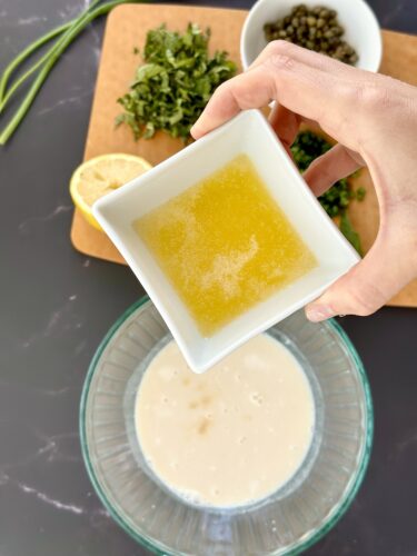 A hand holds a small white bowl of yellow liquid over a glass mixing bowl containing a creamy mixture, with chopped herbs, lemon slices, and capers in the background.