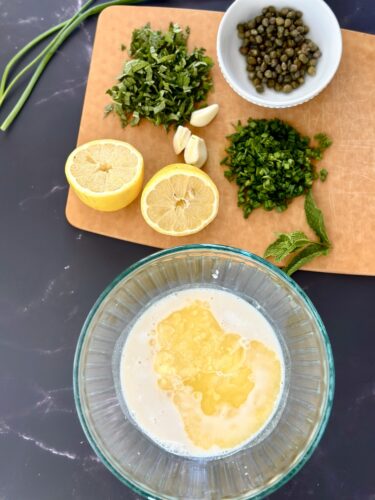 A glass bowl containing a creamy mixture with diced ingredients, surrounded by fresh herbs, garlic, lemon halves, and a bowl of capers on a wooden cutting board.