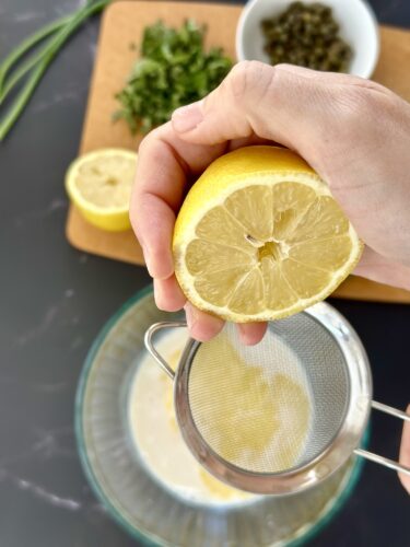 A hand squeezing half a lemon into a strainer over a bowl, with chopped herbs and capers in the background.