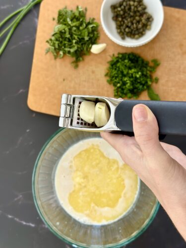 A hand holding a garlic press with two cloves of garlic above a bowl of liquid, with chopped herbs and capers in the background on a cutting board.