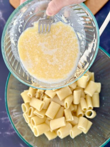 A bowl of melted butter being mixed with a fork, above a glass dish filled with cooked rigatoni pasta.