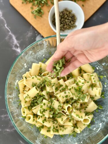 A glass bowl filled with cooked rigatoni pasta mixed with chopped herbs, and a hand adding more herbs to the dish. In the background, there is a cutting board with capers.