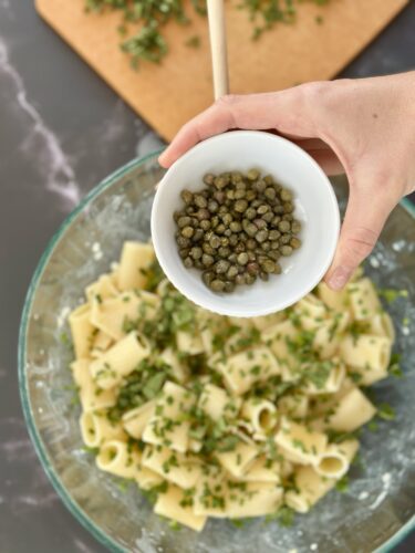 A hand holding a small bowl of capers above a glass bowl of pasta mixed with herbs.