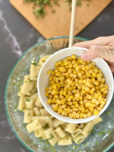 A hand holding a bowl of sweet corn over a large bowl of pasta, with chopped herbs in the background.
