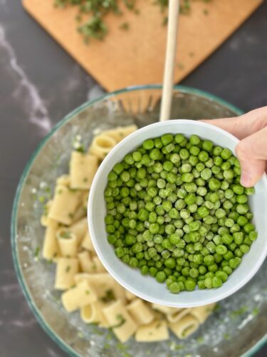A hand holding a bowl of green peas above a bowl of pasta in a glass mixing bowl, with a wooden spoon and a cutting board in the background.